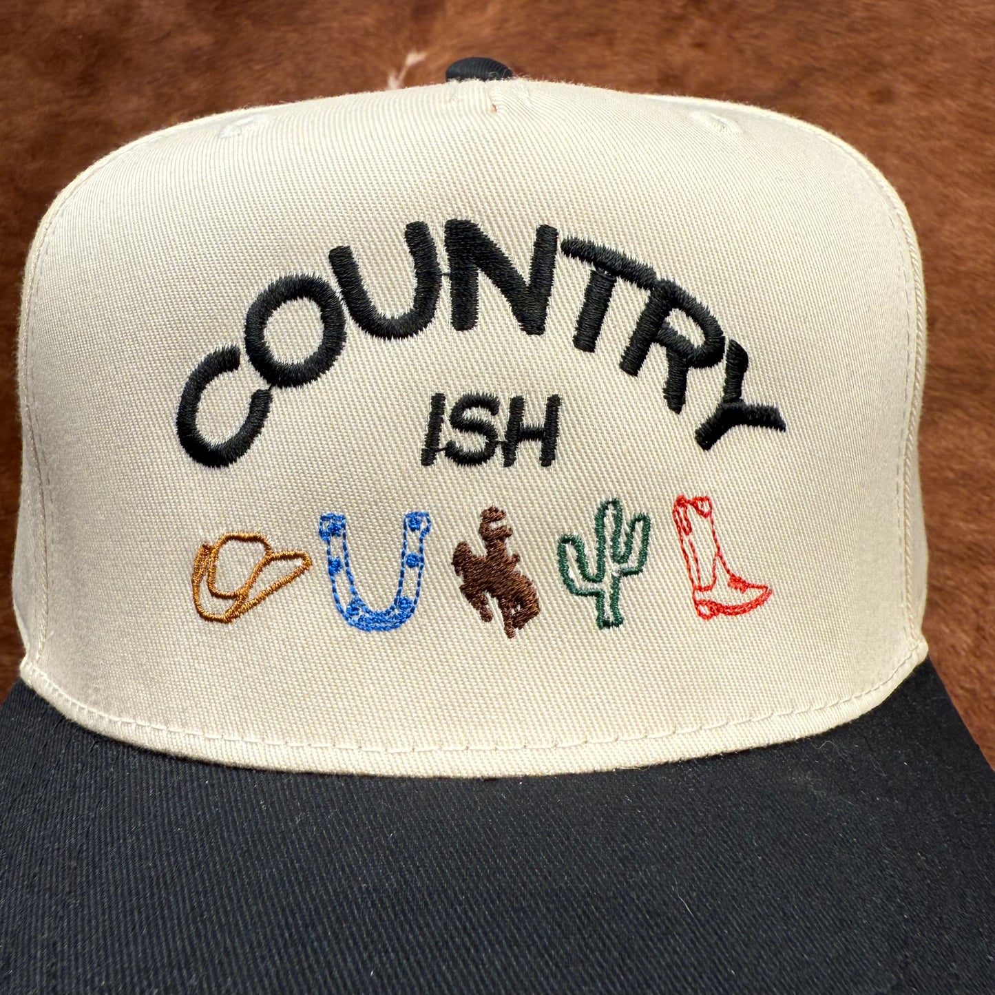 Embroidered western themed trucker hat in Black and beige says "Country ish" with different western themed logos- cowboy hat, horseshoe, buckin' bronc, cactus, and a cowboy boot. Western fashion. Cowgirl outfits. Women's Trucker Hats.