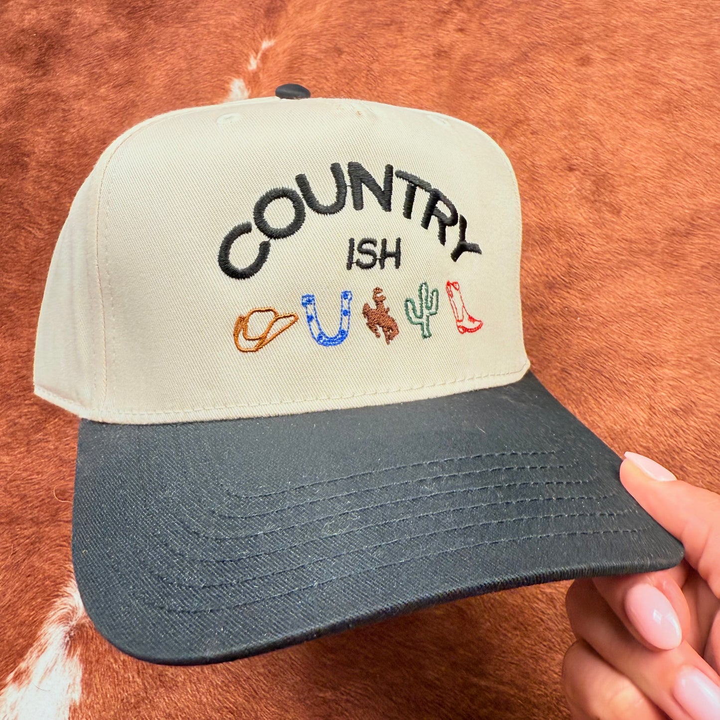 Embroidered western themed trucker hat in Black and beige says "Country ish" with different western themed logos- cowboy hat, horseshoe, buckin' bronc, cactus, and a cowboy boot. Western fashion. Cowgirl outfits. Women's Trucker Hats.