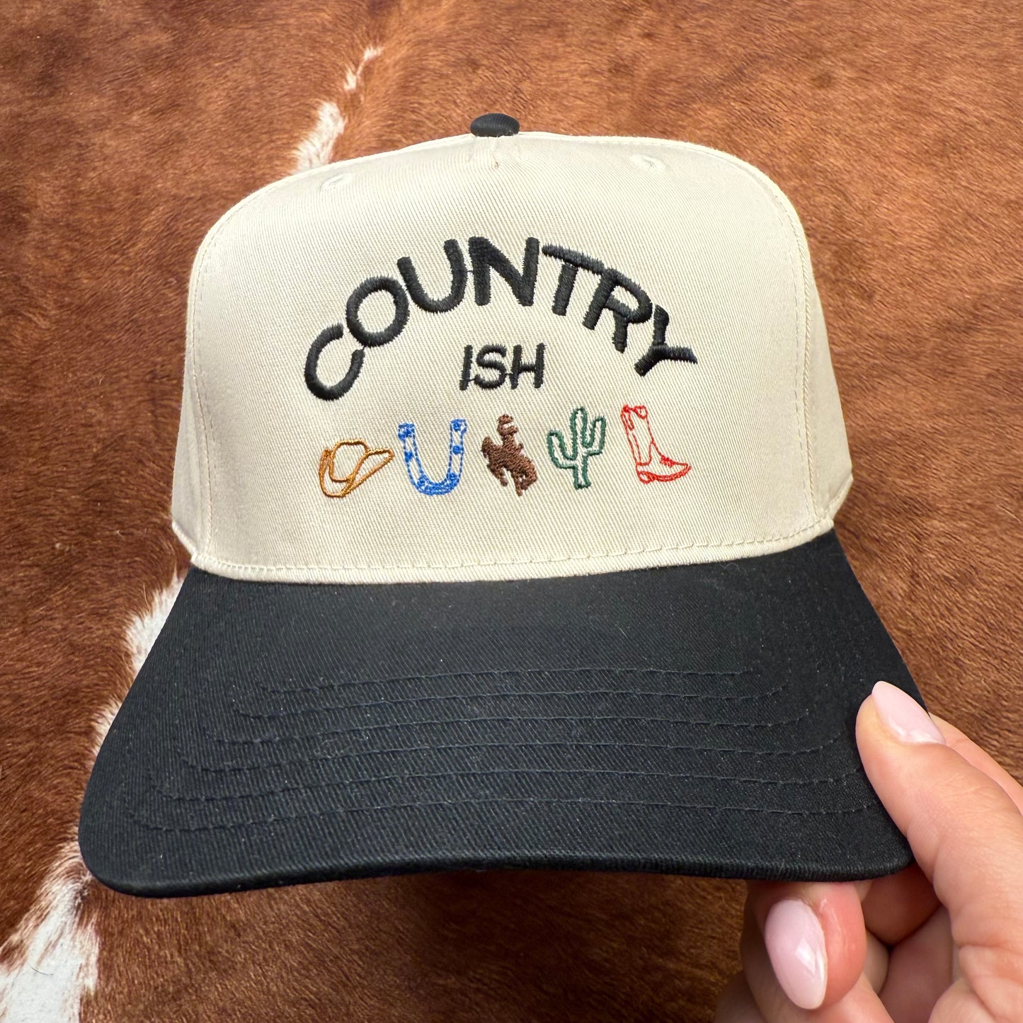Embroidered western themed trucker hat in Black and beige says "Country ish" with different western themed logos- cowboy hat, horseshoe, buckin' bronc, cactus, and a cowboy boot. Western fashion. Cowgirl outfits. Women's Trucker Hats.