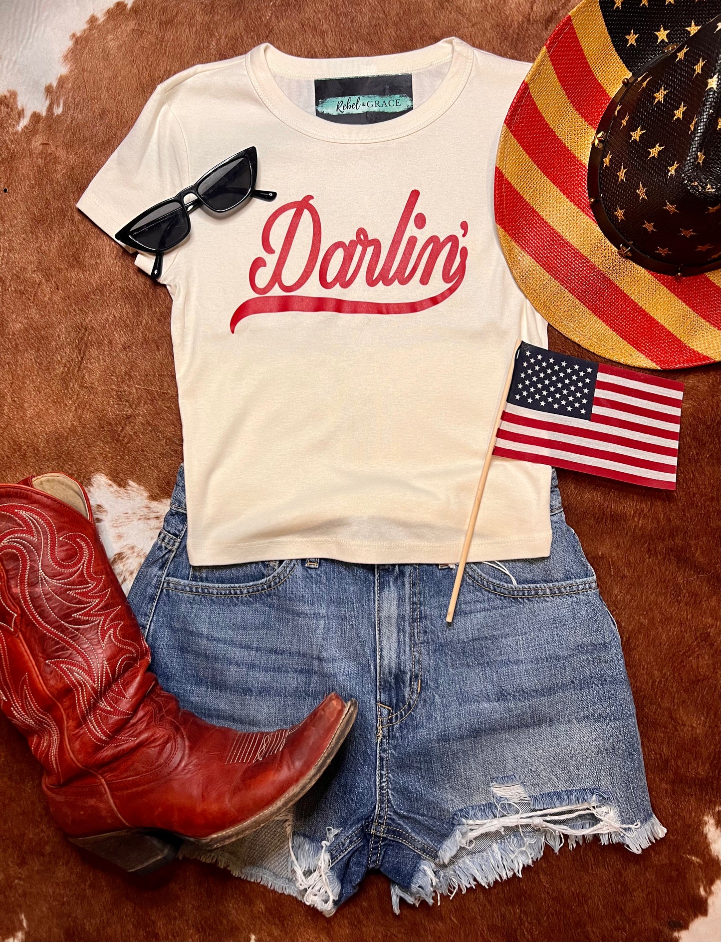 Fourth of July outfit ideas shown is the product a white cropped short sleeve top with Darlin on it in red with a pair of blue denim shorts and a red cowboy boot with american flag cowboy hat and black sunglasses to complete the outfit