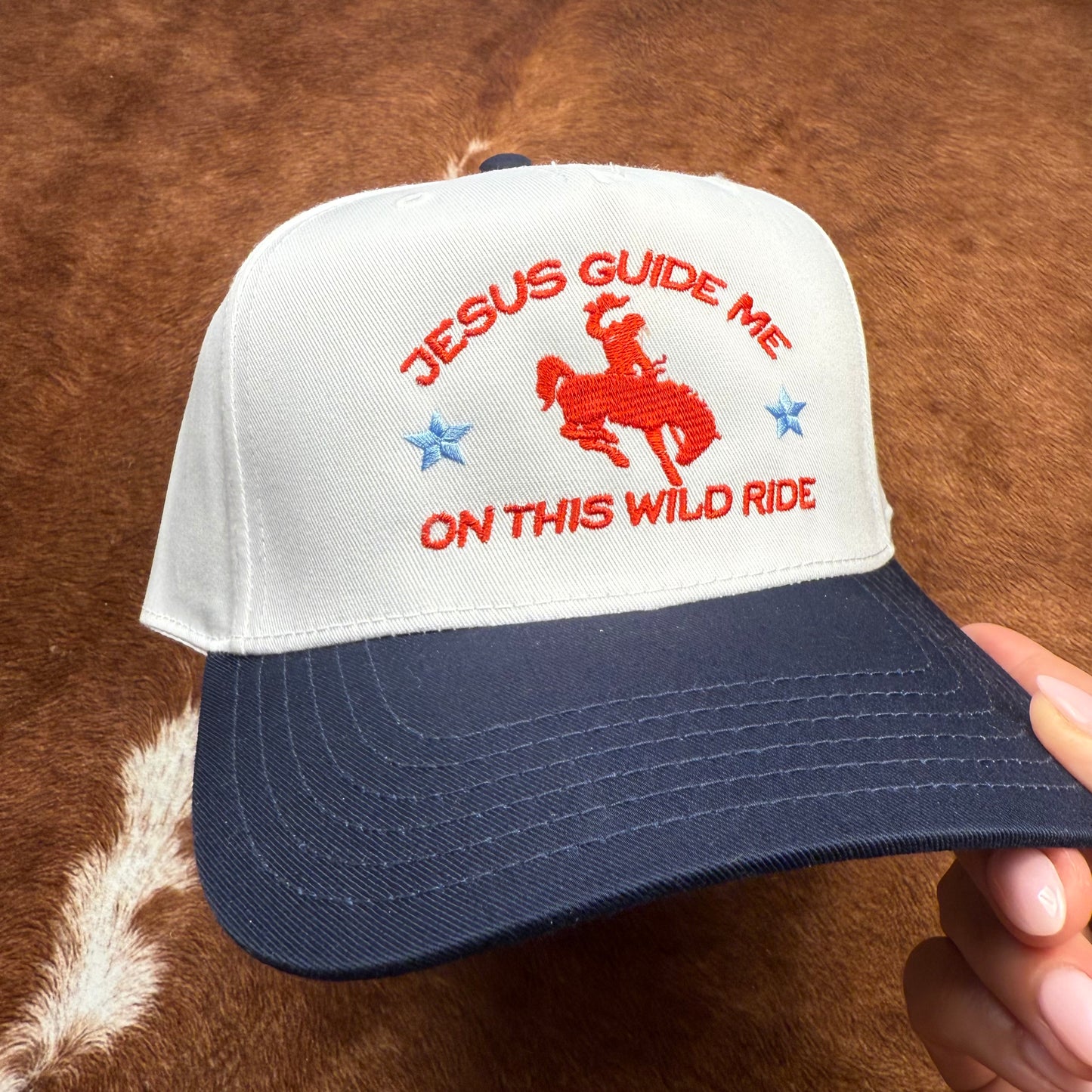 Embroidered western themed trucker hat in navy and white says "Jesus Guide me on this wild ride" with a buckin' bronco with cowboy on it. Western fashion. Cowgirl outfits. Patriotic hats. Christian hats. Trendy Christian fashion.