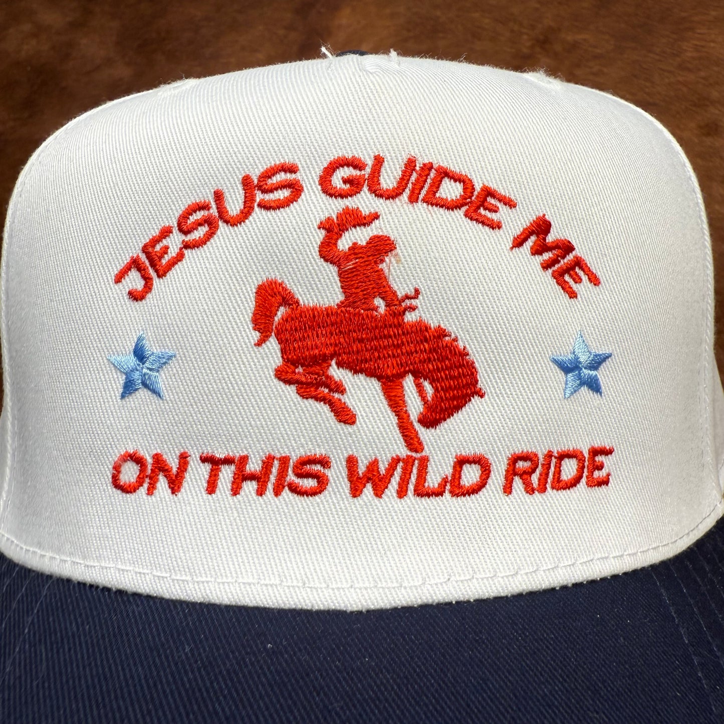 Embroidered western themed trucker hat in navy and white says "Jesus Guide me on this wild ride" with a buckin' bronco with cowboy on it. Western fashion. Cowgirl outfits. Patriotic hats. Christian hats. Trendy Christian fashion.