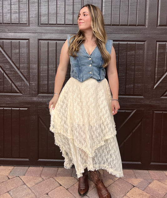 Women in denim button down short sleeve vest. Wearing handkerchief style cream colored lace layered skirt. Cream floral lace tiered ruffled skirt with brown cowboy boots western style festival outfit